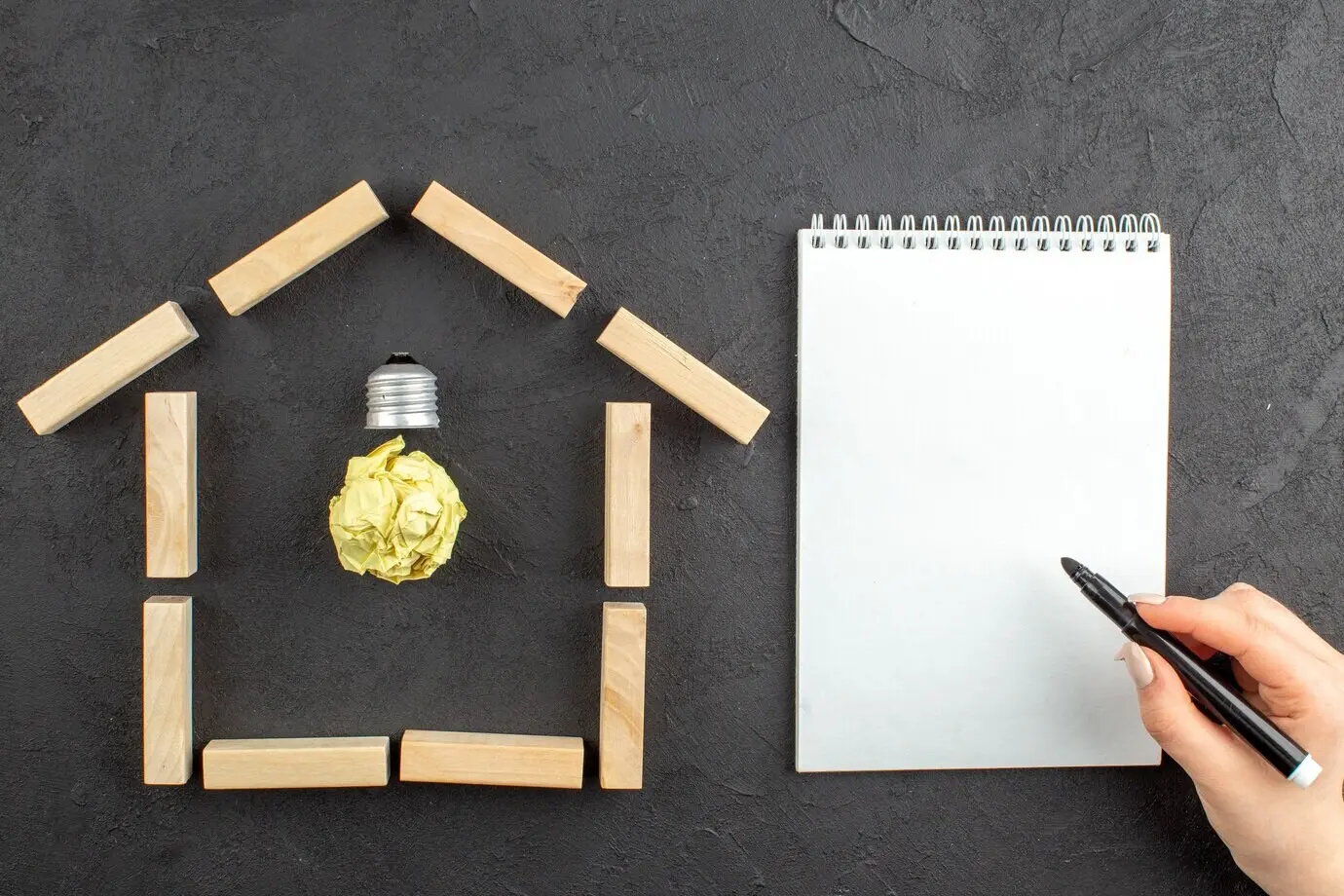 Top view of an idea light bulb, house-shaped wooden blocks, a notepad, and a black marker held in a female hand on a black background.